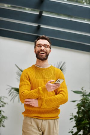 cheerful handsome architect with glasses in yellow turtleneck posing with his phone in his office