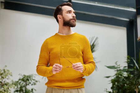 concentrated attractive bearded man in yellow turtleneck holding his glasses and looking away