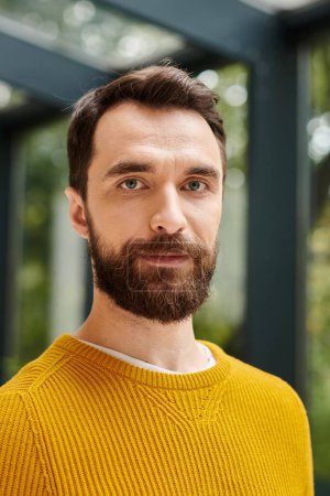 concentrated attractive bearded man in yellow turtleneck looking at camera while in office