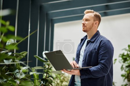 konzentrierter attraktiver Mann in bequemer Alltagskleidung, der seinen Laptop hält, während er hart arbeitet