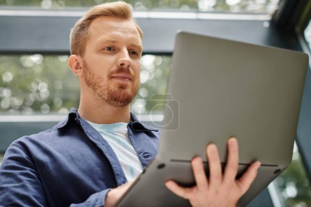 fokussierter gut aussehender Mann in bequemer Alltagskleidung, der seinen Laptop hält, während er hart im Büro arbeitet