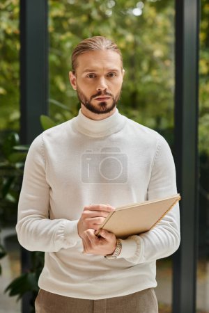 Foto de Hombre atractivo enfocado en blanco cuello alto cómodo con barba sosteniendo su papeleo en la puesta en marcha - Imagen libre de derechos