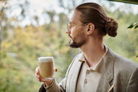 guapo atractivo hombre con barba y pelo recogido en traje elegante bebiendo su café caliente