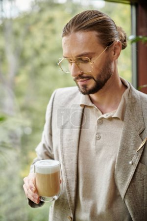 attrayant homme attrayant avec barbe et cheveux recueillis en costume élégant boire son café chaud