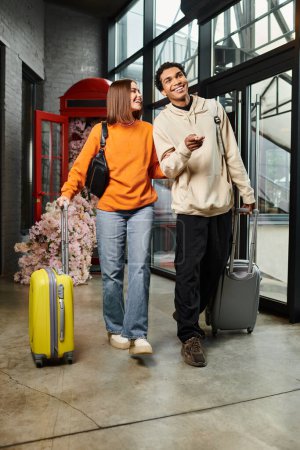 Jeune couple diversifié entrant joyeusement dans un bâtiment moderne avec leurs valises roulantes, auberge