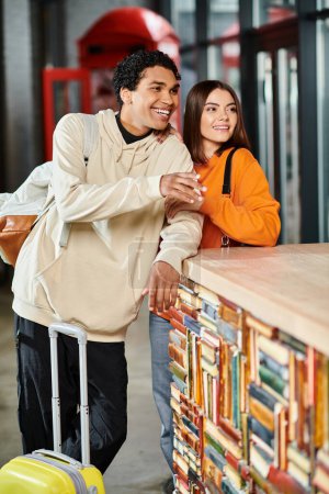 Young couple with luggage leaning on a reception counter, glancing out together in hostel