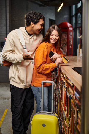 homme afro-américain parlant à une femme heureuse tenant une carte de crédit au comptoir de réception dans l'auberge
