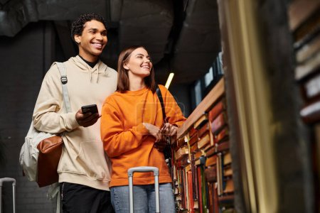 glückliches und vielfältiges Paar mit Smartphone, das einen freudigen Moment in einem Hostel in der Nähe eines Bücherregals teilt
