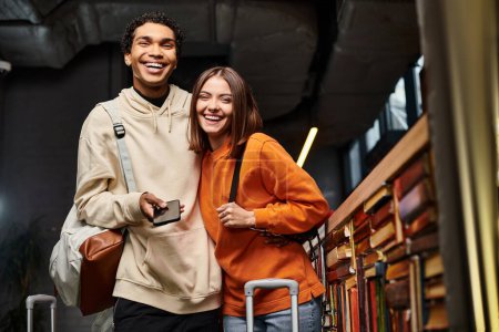 Entzücktes und vielfältiges Paar mit Smartphone teilt einen freudigen Moment im Hostel in der Nähe eines Bücherregals