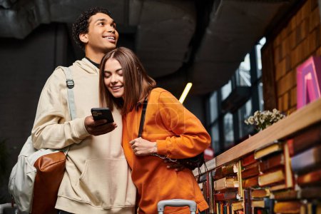 pareja interracial mirando de cerca una pantalla de teléfono inteligente en una biblioteca de albergue, rodeado de libros