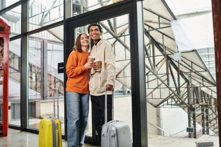 Young diverse couple with travel luggage smiling and entering a modern hostel, holding coffee to go