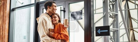 glücklich vielfältiges Reisepaar mit Kaffee umarmt neben modernem Glaseingang der Herberge, Banner