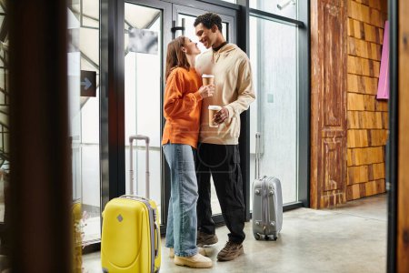heureux couple interracial avec café embrassant à côté des bagages de voyage dans une entrée moderne de l'auberge