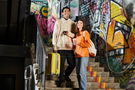 feliz pareja diversa de pie en las escaleras con graffiti en la pared, hombre negro sosteniendo una maleta amarilla