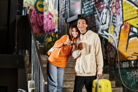 diverse couple smiling and standing next to each other on stairs with graffiti, man holding luggage