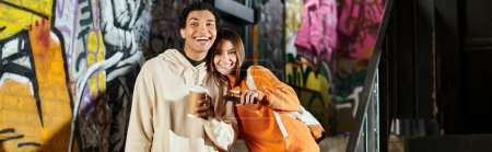 Couple heureux en tenue décontractée avec tasse à café et smartphone près du mur peint graffiti, bannière