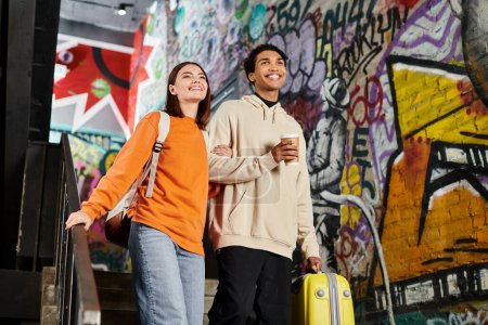 couple diversifié souriant et marchant l'un à côté de l'autre dans les escaliers avec graffitis, café et bagages