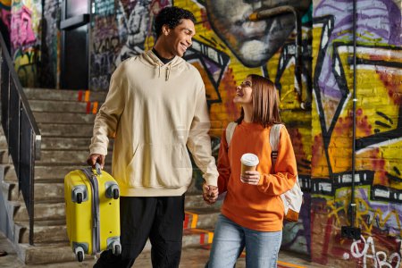 pareja diversa tomados de la mano y sonriendo con una maleta amarilla en una pared pintada de graffiti, albergue