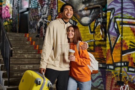 happy diverse couple embracing by a staircase with graffiti on background, black man with luggage