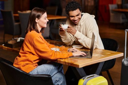 Happy woman showing something on laptop to black man while he holding coffee cup in cafe, diversity