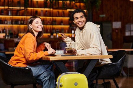 diverse couple having coffee break and doing online shopping near luggage in cafe, credit card