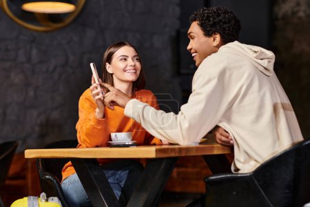 Téléchargez les photos : Femme heureuse en pull orange montre téléphone au petit ami noir souriant tout en étant assis dans le café - en image libre de droit