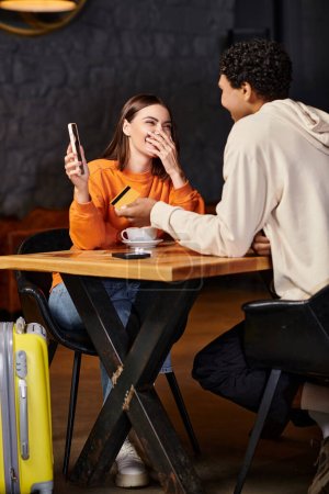 Kobieta zakrywa usta śmiejąc się kiedy jej czarny chłopak szczęśliwie rozmawia z nią w przytulnej kawiarni