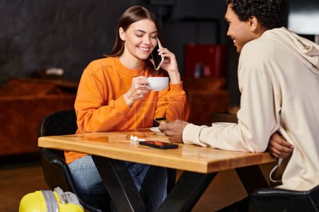 Sonriente joven chateando en su teléfono inteligente con una taza de café cerca de novio negro en la cafetería