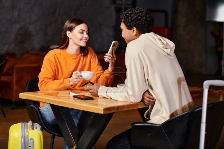 Smiling young woman using her smartphone and holding coffee cup near black boyfriend in cafe