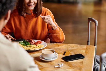 happy woman eating breakfast near warm cup of coffee while enjoys tasty meal near black boyfriend