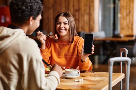 happy young woman in orange sweater happily showing her phone to black boyfriend during meal