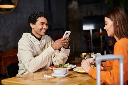 glücklicher schwarzer Mann mit Smartphone, während ihm eine Frau beim Frühstück gegenübersitzt
