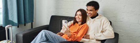 heureux couple diversifié partage un moment confortable, défilant à travers les téléphones sur un canapé confortable, bannière