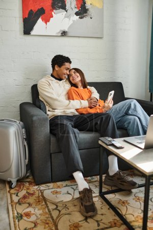 Photo for Happy diverse couple cozily lounges on a couch in their stylish living room surrounded by gadgets - Royalty Free Image