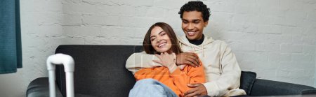 Photo for Happy multiethnic couple cozily hugging on a couch in stylish living room of hostel, banner - Royalty Free Image