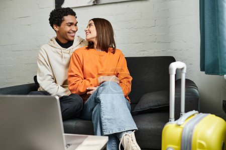 pareja multiétnica positiva relajarse en un sofá en su elegante sala de estar cerca de la computadora portátil en la mesa