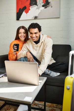 feliz pareja diversa comparte un momento acogedor mientras se sientan en el sofá y el uso de la computadora portátil, taza de café