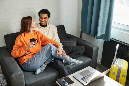 Photo for Happy diverse couple in cozy clothing sitting on a couch in hostel, surrounded by laptop and luggage - Royalty Free Image