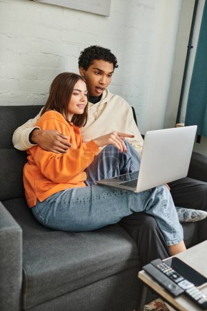 Photo for Diverse couple sitting on couch and browsing laptop, woman pointing at screen near black boyfriend - Royalty Free Image