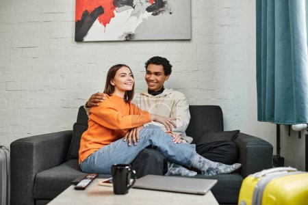 cheerful diverse couple enjoys cozy moment on a sofa in hostel, with travel bags indicating a trip
