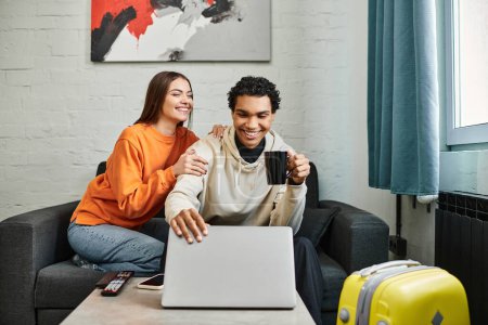 Excited diverse couple seated on couch with a laptop, possibly booking a trip in hostel room