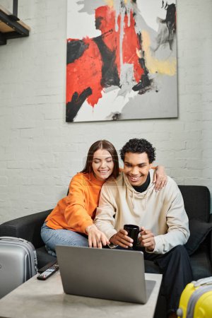 Téléchargez les photos : Couple souriant et diversifié assis confortablement sur un canapé, posant avec un ordinateur portable dans un espace de vie moderne - en image libre de droit