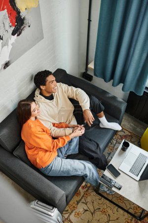 jolly diverse couple enjoys a calm moment while relaxing on sofa with a gadgets and luggage