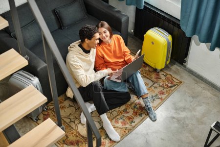 Lächelndes gemischtrassiges Paar sitzt neben Sofa mit Laptop, plant eine Reise oder bespricht Zukunftspläne