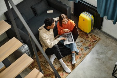 diversa pareja mirando a un ordenador portátil mientras planea su próximo viaje, sentado en la alfombra cerca de las escaleras