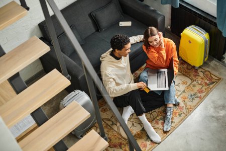 multiethnic couple looking at a laptop while planning their next trip, sitting on carpet near stairs