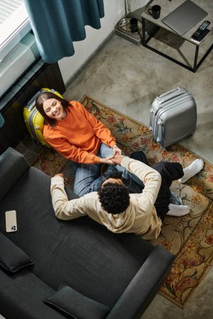 vista superior de una pareja relajada y diversa sentada en el suelo con un ordenador portátil, disfrutando de un acogedor día en el interior