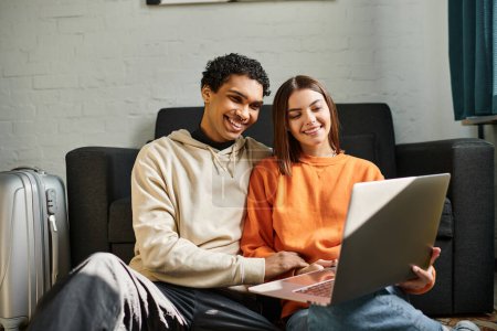 Smiling multiethnic couple comfortably using a laptop together on a dark sofa, couple goals