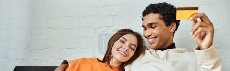 Cheerful diverse couple looking at a credit card while cozily reclining on living room sofa, banner