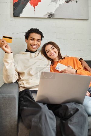 glückliches multiethnisches Paar, das online einkauft, eine Kreditkarte in der Hand, mit einem Laptop auf dem Sofa im Hostel